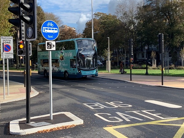 "The words 'bus gate' are painted on the road. To the left of the road markings there's a blue, circular signpost next to a set of traffic lights, the sign shows a bicycle, bus and the word taxi"