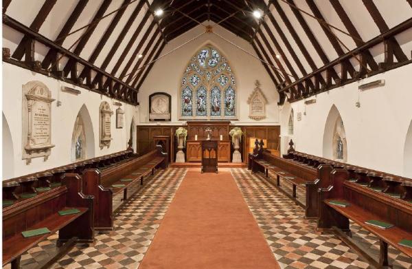"Inside view of the Extra-Mural chapel. a wide aisle leads to a lecturn in front of a stained glass window. Wooden pews face into the aisle on either side."