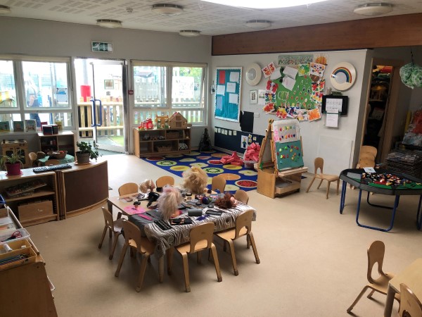 "play room with open door to outside play area. There are tables with toys and activities and art on the walls."