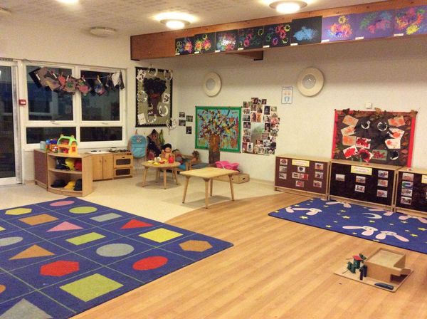 "play room with a large rug with shapes on the floor. there are tables with toys and children's artwork on the wall"