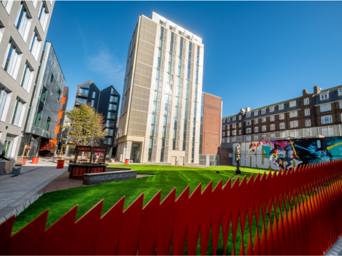 Looking over a rusty-re coloured metal railing with a series of teeth-like triangular tops you can see a lawn leading to a brand new tall building. The building is predominantly white with a lot of windows, and stands out againsta bright blue sky. To the left is another new building.