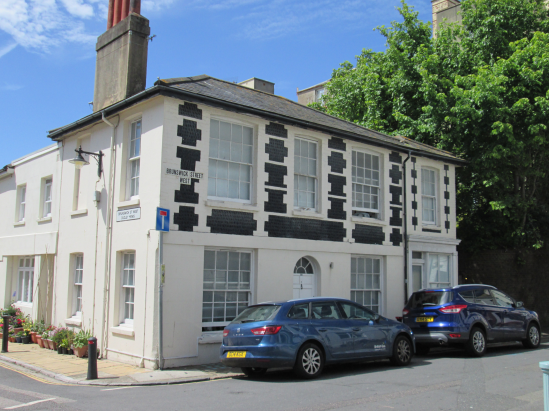 A large 2 storey builidg with large windows. There is a black brickwork pattern on the upper frontage of the otherwise white house