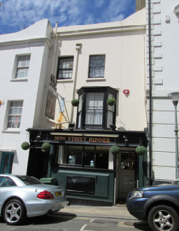 The front of the Three storey building containing the Bow Street Runner pub. The ground floor is the black and dark green pub frontage, with name and swinging sign. Above it is a black bay window. THe rest of the building is white