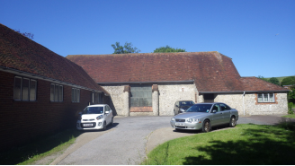 A converted barn with long slopig brown roofs and gret stone walls. It sits in an L-shape around a courtyard where two cars are parked.