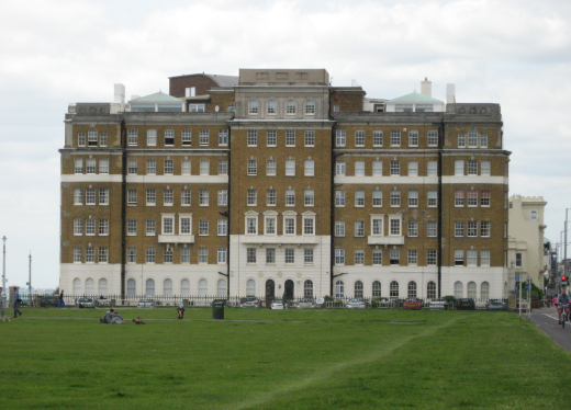 A large building on the seafront at the end of a stretch of lawn. It is 8 storeys high and 22 windws wide. The first floor brick is white, the roof is grey and the rest f the building is a sandy brown. 