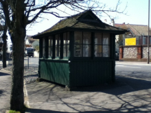 A small, standalone green wooden shelter. It has a grey wooden roof and windows on three sides. The other side contains three open entranceways