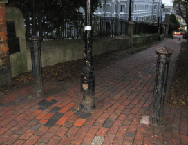 Two black cast iron bollards, about waist height, are fixed either side of a black lampost. There are set into a pedestrianised, redbrick cobbled alleyway.