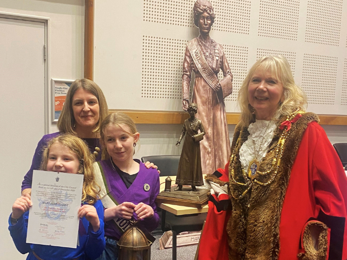 The image shows the Mayor of Brighton & Hove, Councillor Jackie O'Quinn, wearing her ceremonial robes standing next to a blonde woman, Briony Goulden, and her two daughters, dressed in purple, sporting 'Votes for Women' sashes and holding the Freedom of the City certificate. Behind them there is a life sized cardboard cut out of Mary Clarke and a miniature statue of her. 