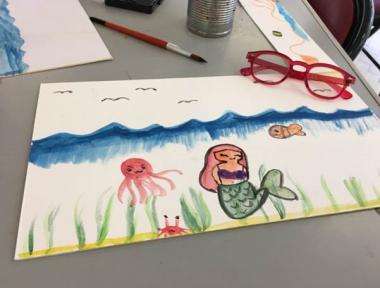 Painting of a mermaid, octopus, crab and fish on a white bit of paper. There are red glasses on top of the corner of the painting.