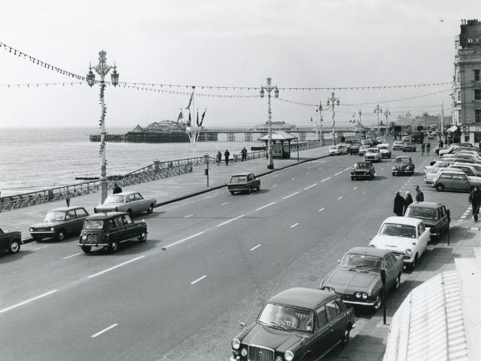 An historic photo of a seafront road with streetlights, cars and a pier in the background