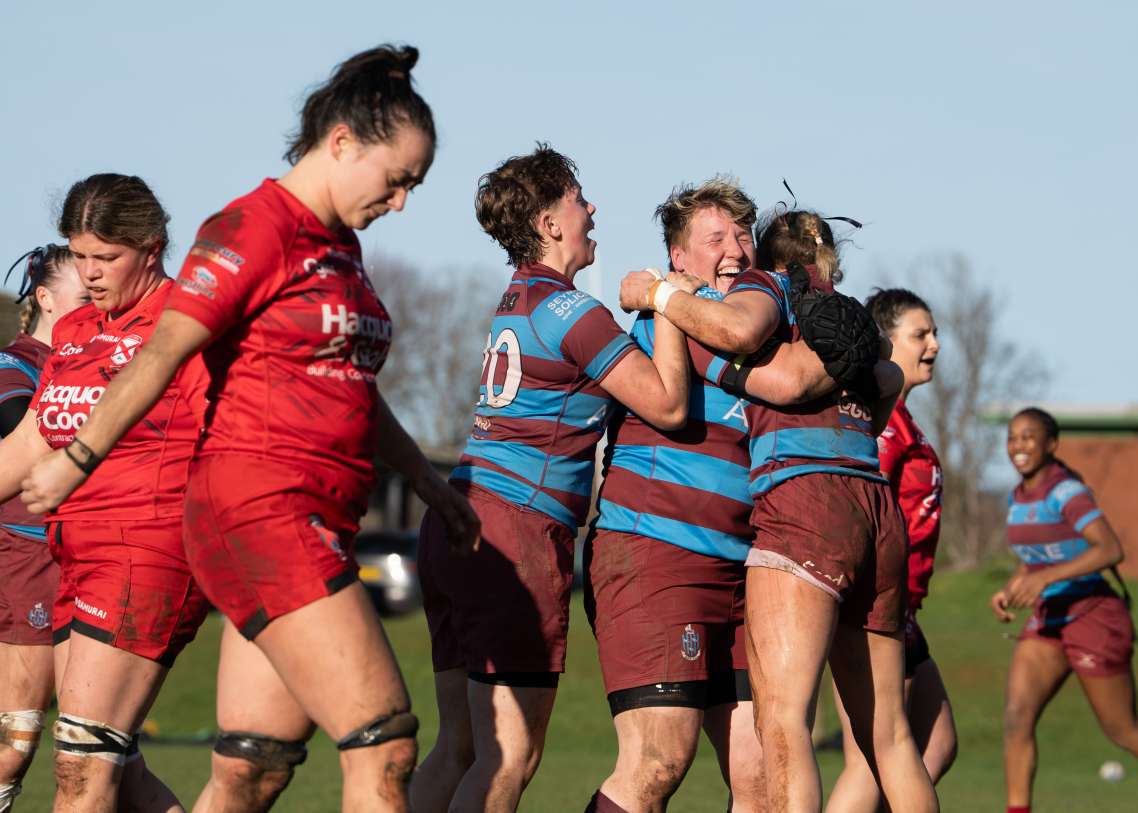Three players in maroon and blue kit celebrate a try together, while opponents in red look dejected