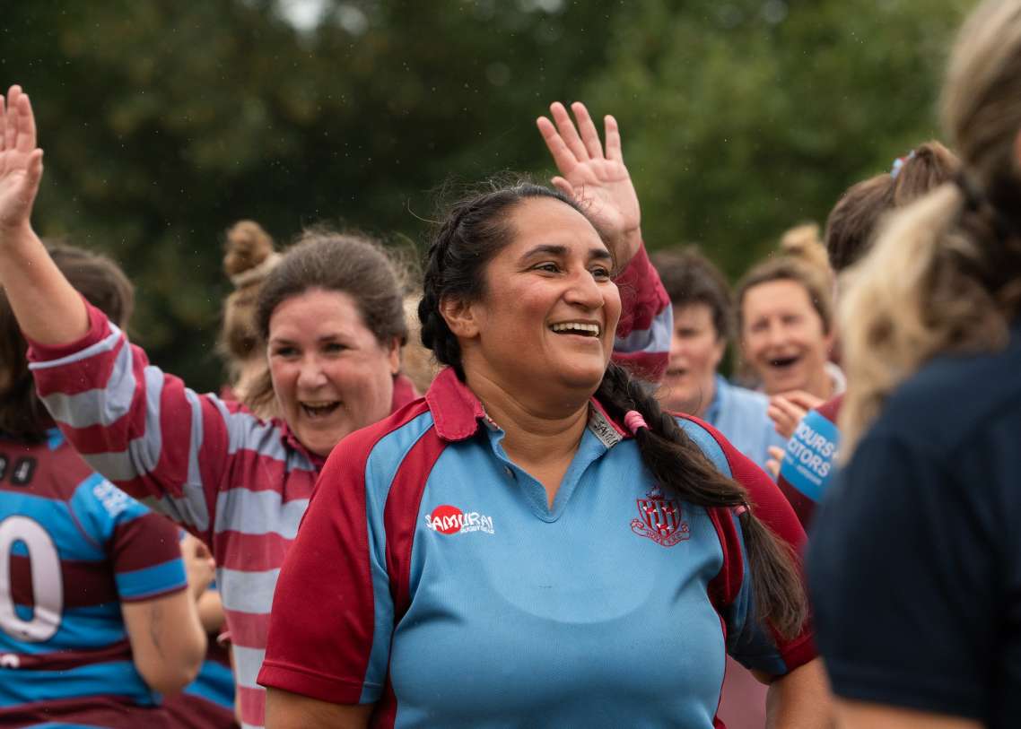 A rugby player in a blue and maroon top smiles, with teammates in the background also smiling