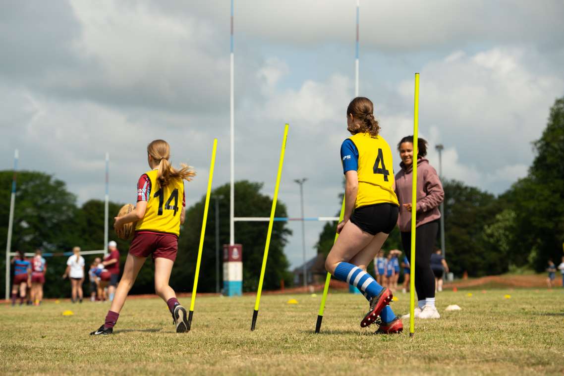 Two players in yellow bibs, one with number 14 on their back and one with number 4 on their back, run between poles being encouraged by a coach