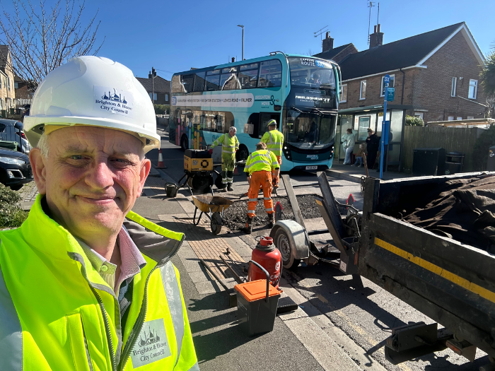 A man in a high viz jacket and hard hat posing in front of a group of men in similar clothing working on a road with a bus in the background
