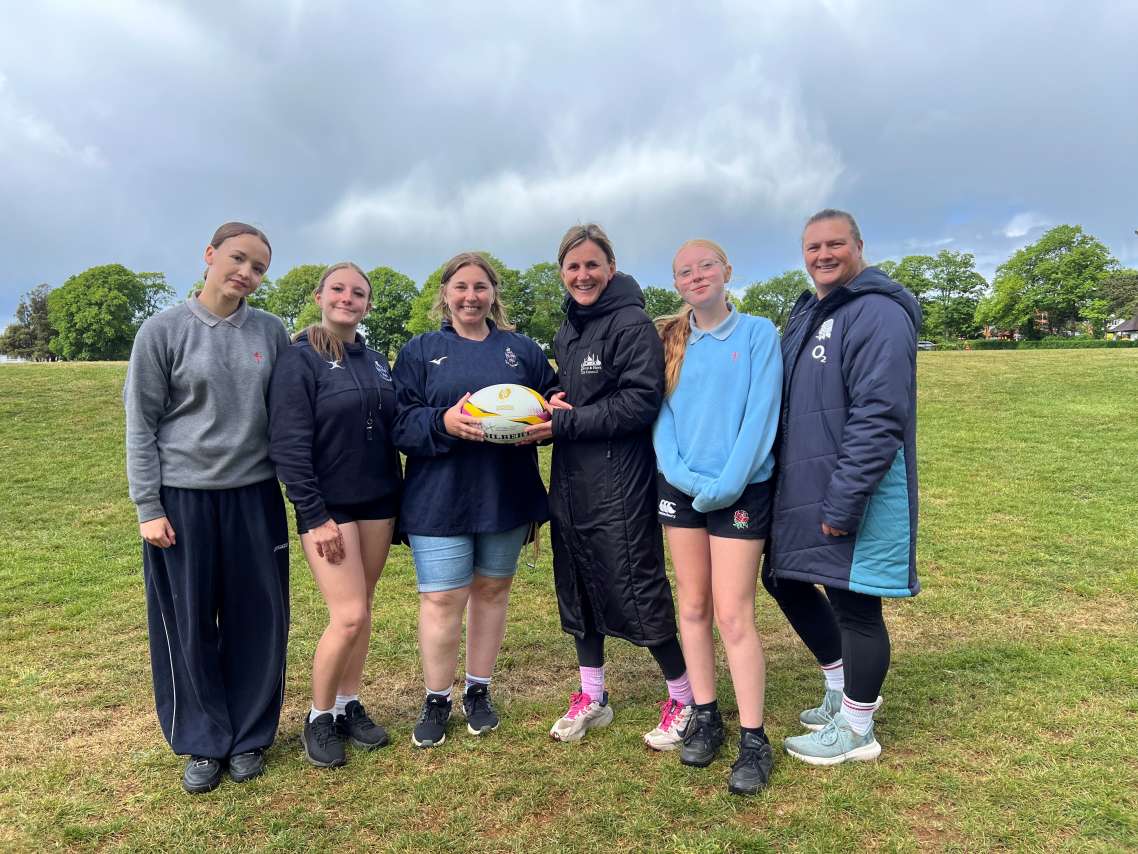 Three teenage girls and three women in a group photo pose, with the two in the middle holding a rugby ball together. All stood on a rugby pitch.