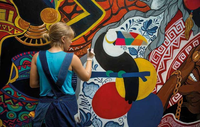 Person painting a Toucan on a wall already decorated with colourful patterns