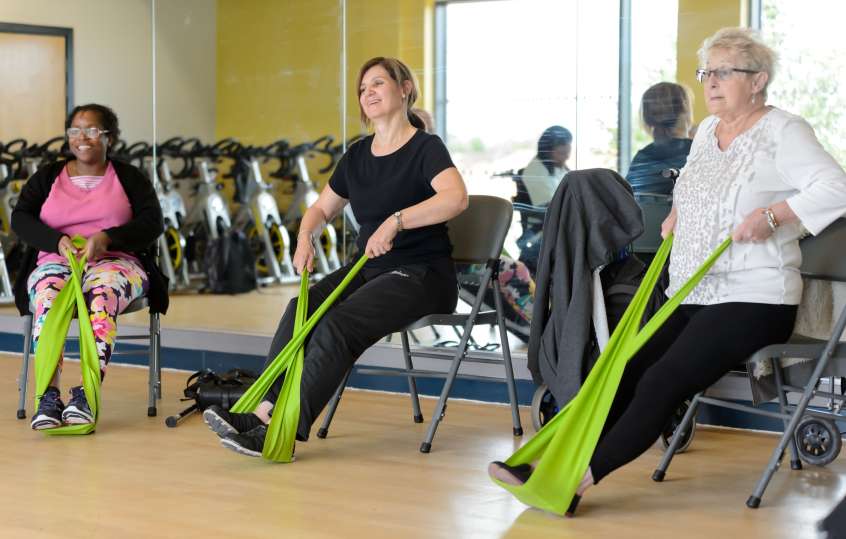 Three women sitting on chairs doing stretching exercises in a gym