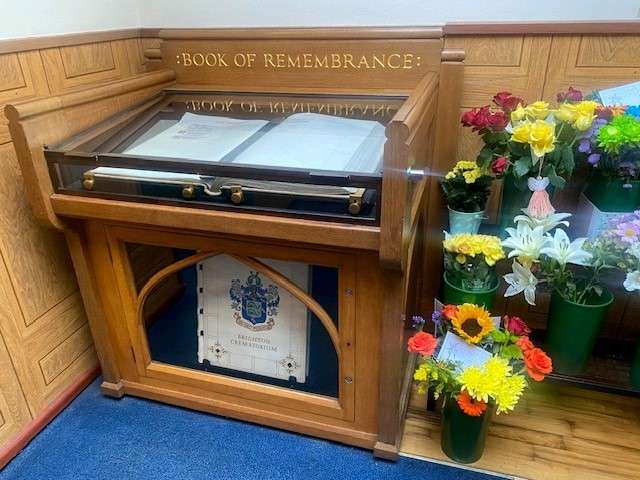 Book of remembrance in a glass case, on a wooden stand. Bouquets of flowers next to the stand.