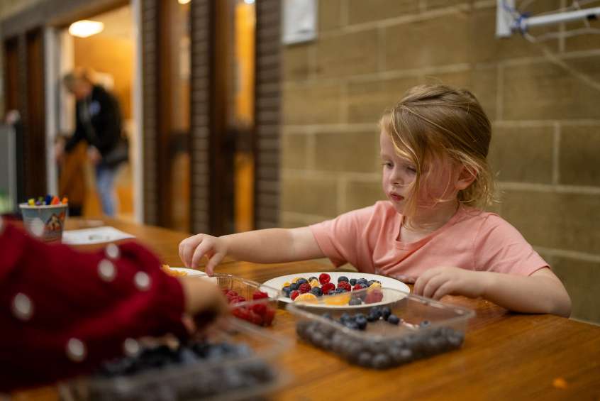 A little girl in a pink t-shirt sits at a table with a plate of fresh fruit.