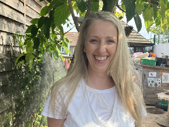 Suzzane standing in front of a wooden wall, tree with leaves with a wall in the background.