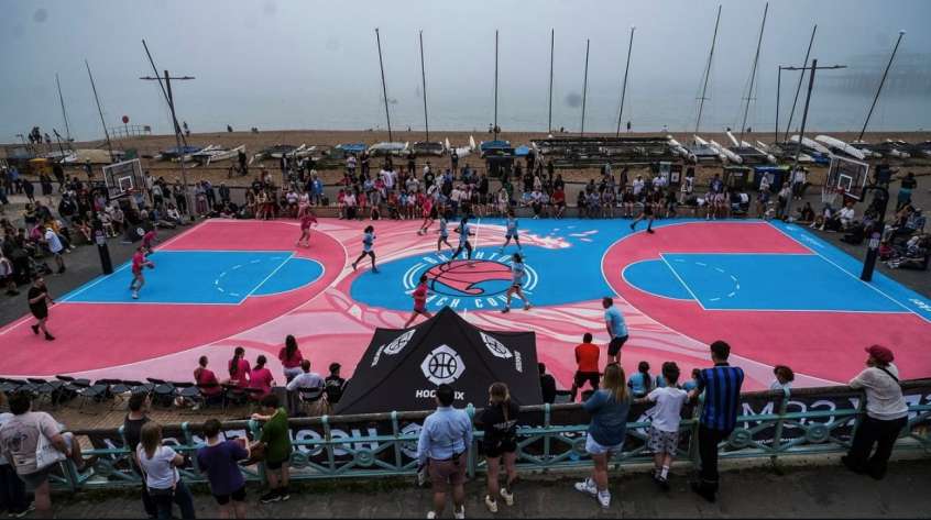 A pink and blue coloured basketball court next to a pebble beach. There is a small crowd standing around the edge watching the players play on the court.