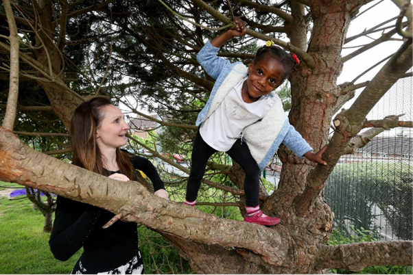 Nursery worker standing next to a child while they play in a tree.