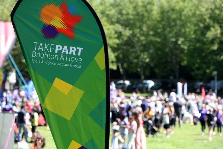 A banner for TakePART festival is in focus in the foreground. Behind it is a big gathering of people in the park.