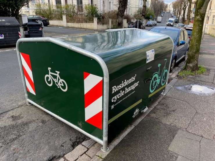 A large green shelter on the side of the street. It has a logo of a bike, and the words 'Cycle Hangar' printed in white. On the corners are large reflective stickers for safety