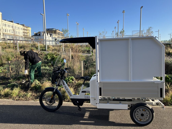 The E-Cargo bike is parked on the verge of a road and a council worker is gardening behind it.