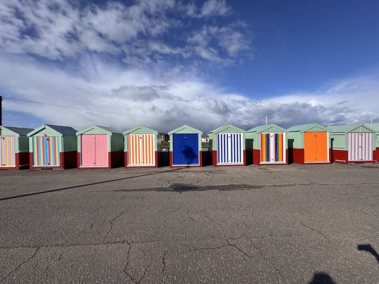 A line of beach huts on Hove Promenade against a blue sky. The top half of each hut is green, the bottom half red. Each hut has it's own distinctive front door - ranging from bright, multi-coloured striped designs, to blocks of vivid blues or oranges.