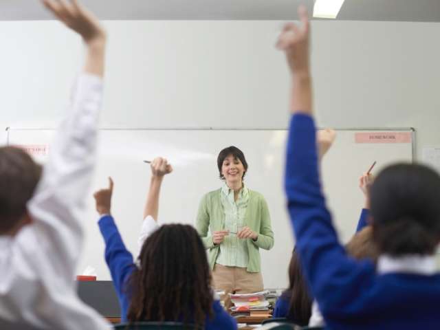 Children raises their hands as a teacher stands at the front of the classroom