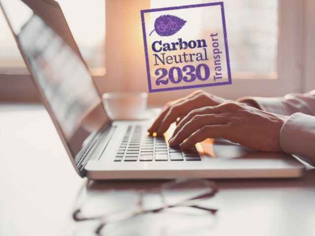 Person's hands typing on a laptop keyboard. The laptop is on the desk. A logo is above the hands with an illustration of a leaf and the words: Carbon Neutral 2030, transport