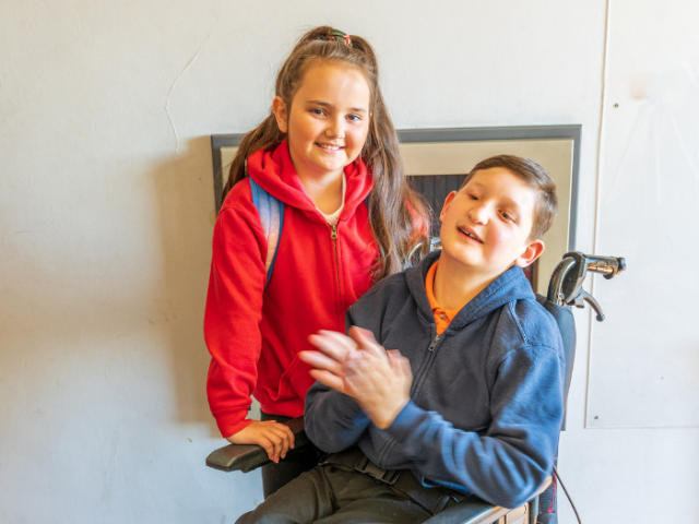 A young girl in a red top with her younger brother in a blue jumper who is in a wheelchair