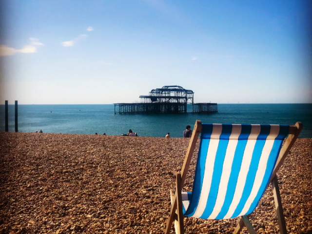 An empty blue and white striped deckchair on the beach with Hove pier in the background
