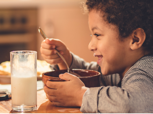Young boy sitting at a table with a glass of milk and a bowl with a spoon eating 