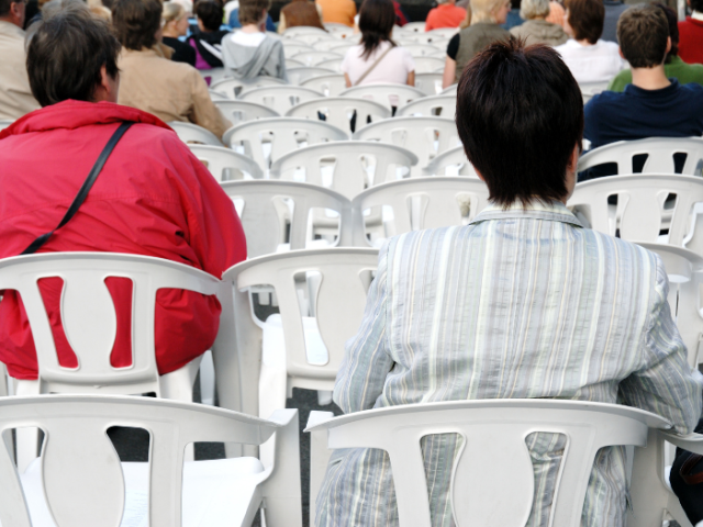 People sitting down watching an outdoor event  on white chairs. View is from behind and people are positioned at a physical distance from each other.
