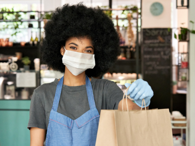 Woman working in a coffee shop wearing a mask and an apron, holding out a paper bag 