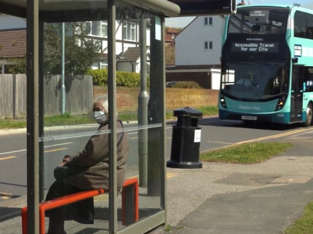 Photo of a woman at a bus stop wearing a mask with a Brighton & Hove Bus in the background