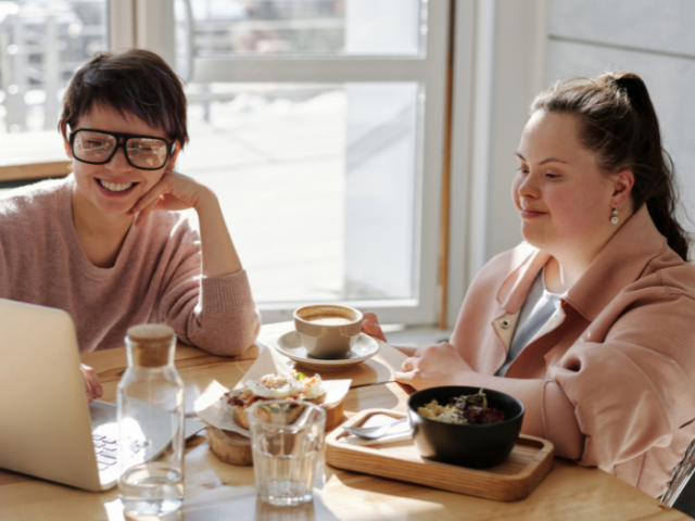 A woman with short dark hair and glasses looking at a laptop and talking to a woman with long dark hair 