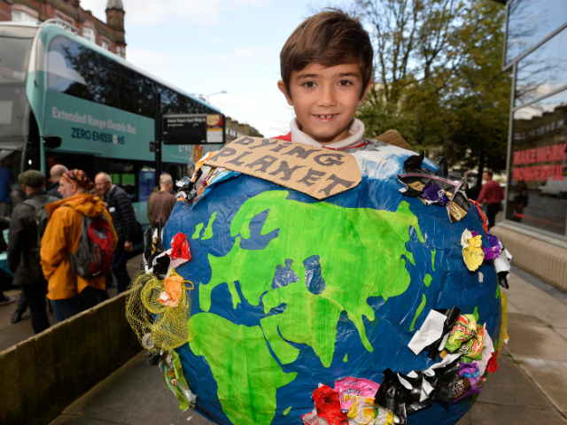 A schoolchild holding a paper cache map of the east with a sign saying 'dying planet' pinned to it. There is a bus and other people gathered in the background.