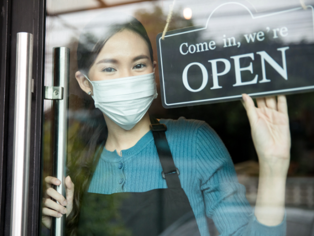 Woman shopkeeper wearing a mask, turning shop door sign to 'open'.