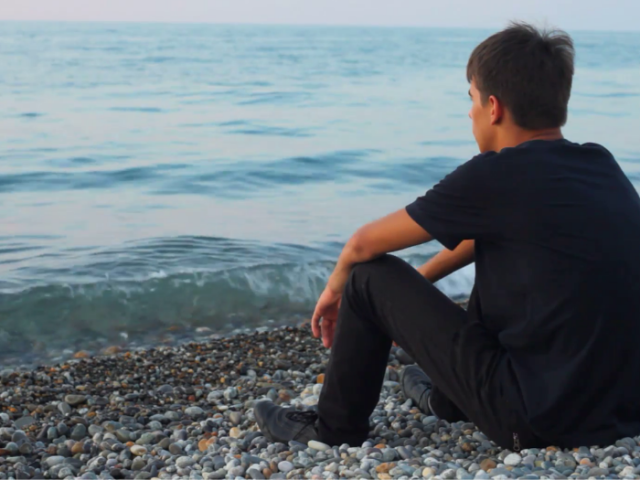 A young man sitting on pebble beach looking out to see