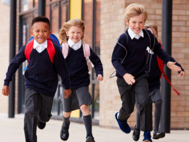 Three children in school uniform with backpacks on running out of school onto a playground, possibly at the end of a school day.
