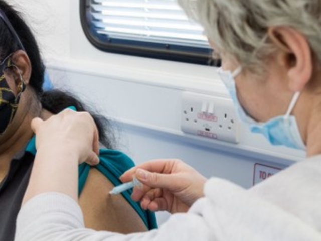 Woman in a white top and wearing a mask is giving a vaccine to another woman in a dark top who is wearing a mask.