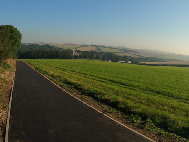 A picture of a black path next to a large green field and blue sky
