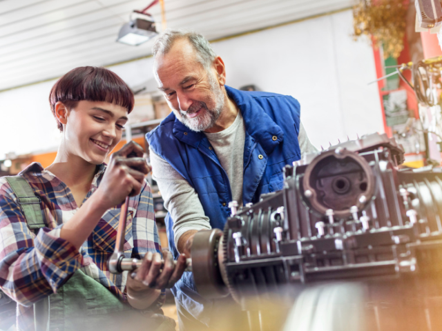 Older man looking over at a young woman fixing an engine 