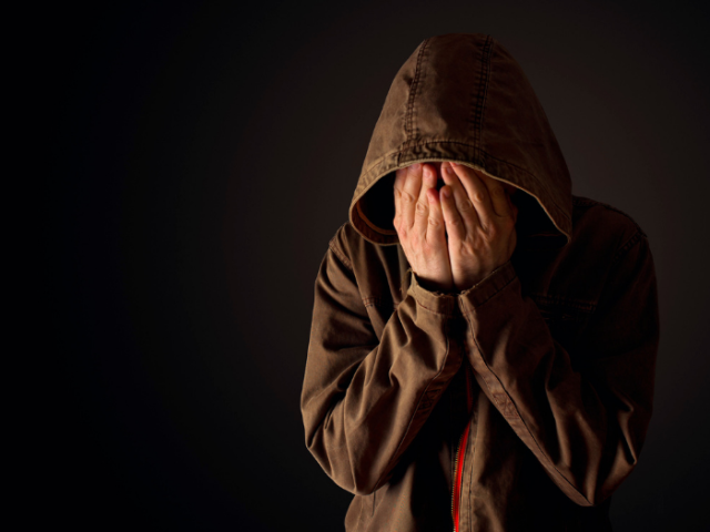 Photo of an anonymous distressed young person in a hooded top holding their face in their hands.