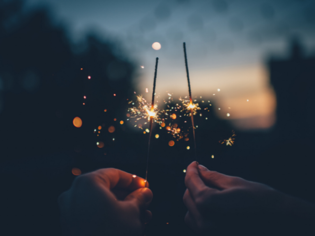 Photo showing hands holding two sparklers with trees and houses silhouetted in the background.