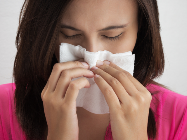 Woman with brown hair and pink top sneezes into a tissue