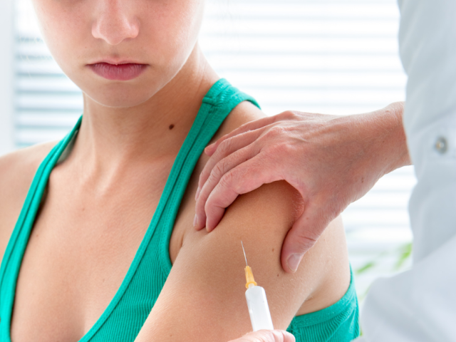 Female teenager receives Covid vaccine from a syringe used on her upper left arm by a person in a medical whitecoat on the right